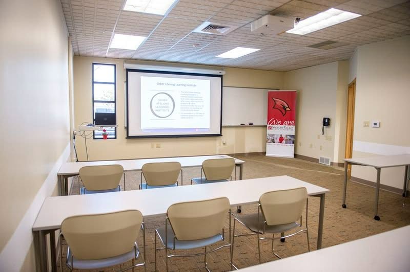 a classroom with rectangular tables and chairs facing a projector screen